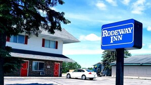 Un letrero de Rodeway Inn frente a un edificio de dos pisos con una puerta roja y un coche blanco estacionado cerca.