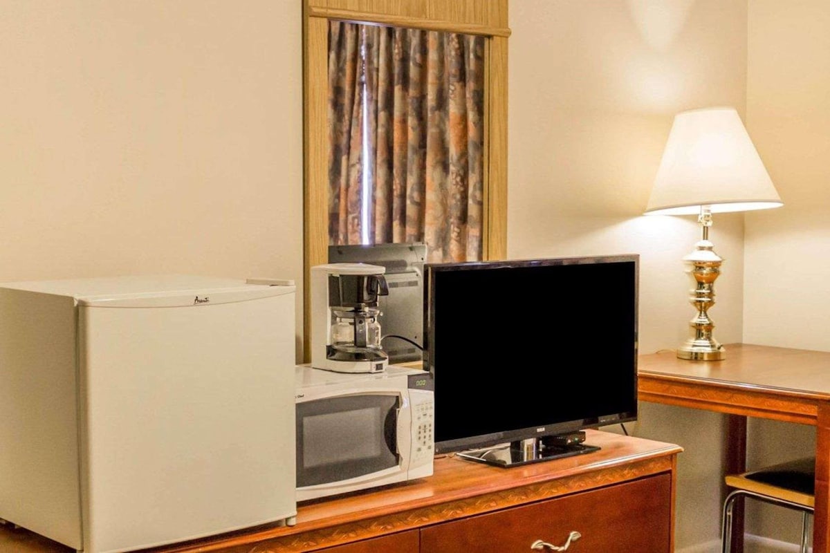 A hotel room with a microwave, a coffee maker, a television on a wooden cabinet, a lamp, and a window with curtains.