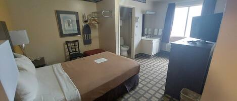 A hotel room with a bed, a desk, a chair, a small table, a TV, a bathroom, and a window.