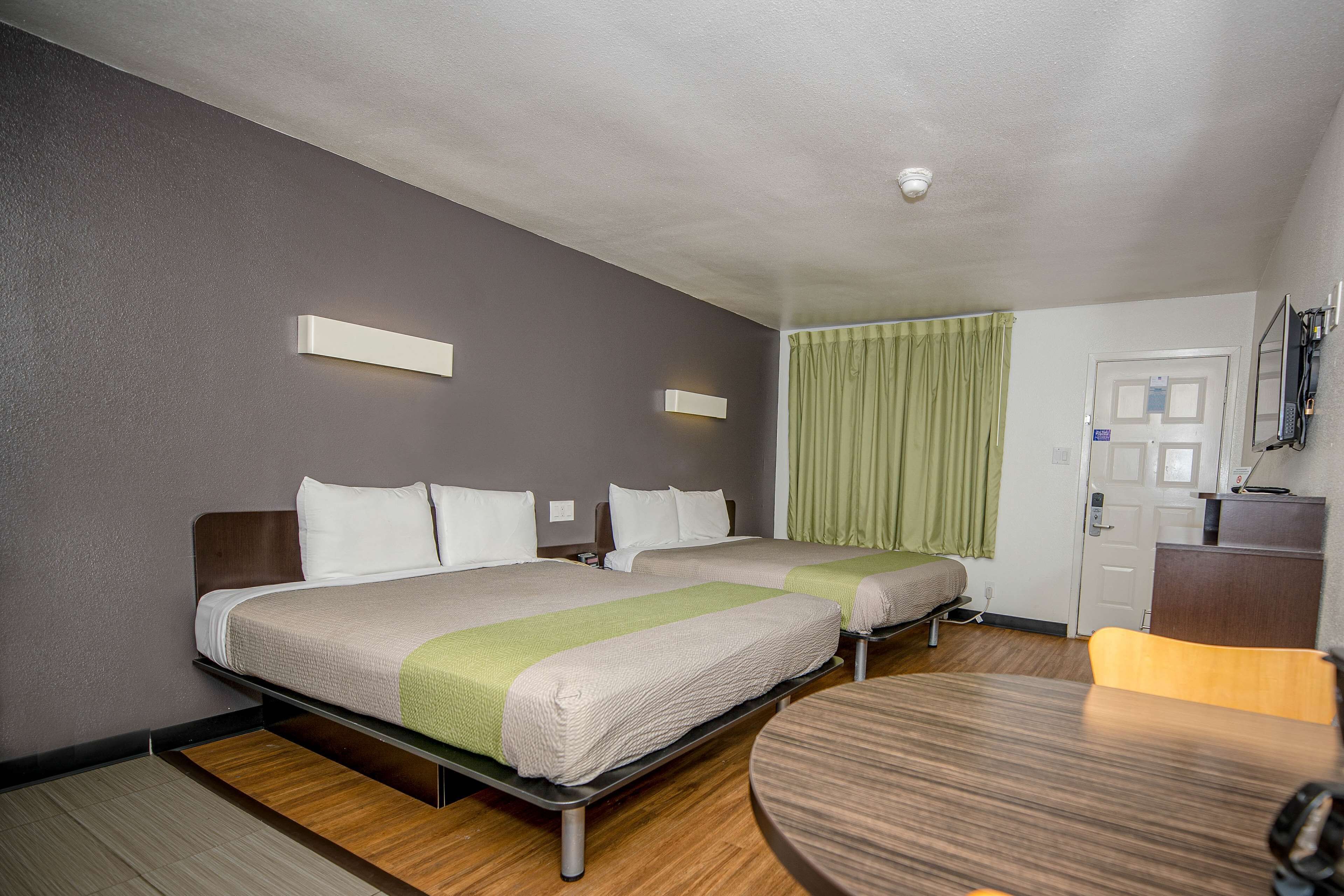 A hotel room with two beds, a desk, and a chair. There is a television on the wall and a door with a lock.