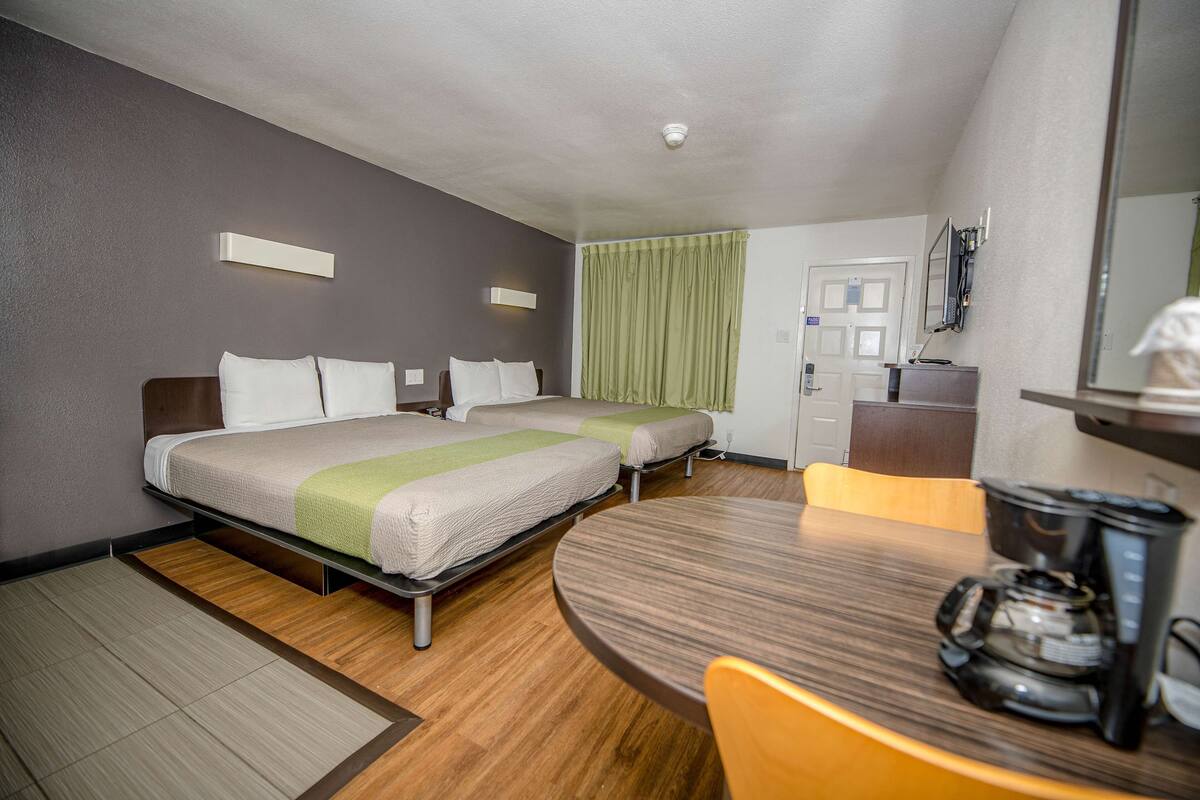 A hotel room with two beds, a dining table, a coffee maker, and a mirror.