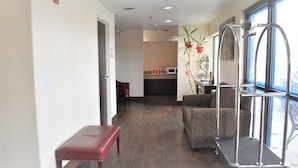 A hotel corridor with a bench, wicker furniture, a cart, and a kitchen area in the background. - Super 8 by Wyndham New Braunfels I-35 (New Braunfels)