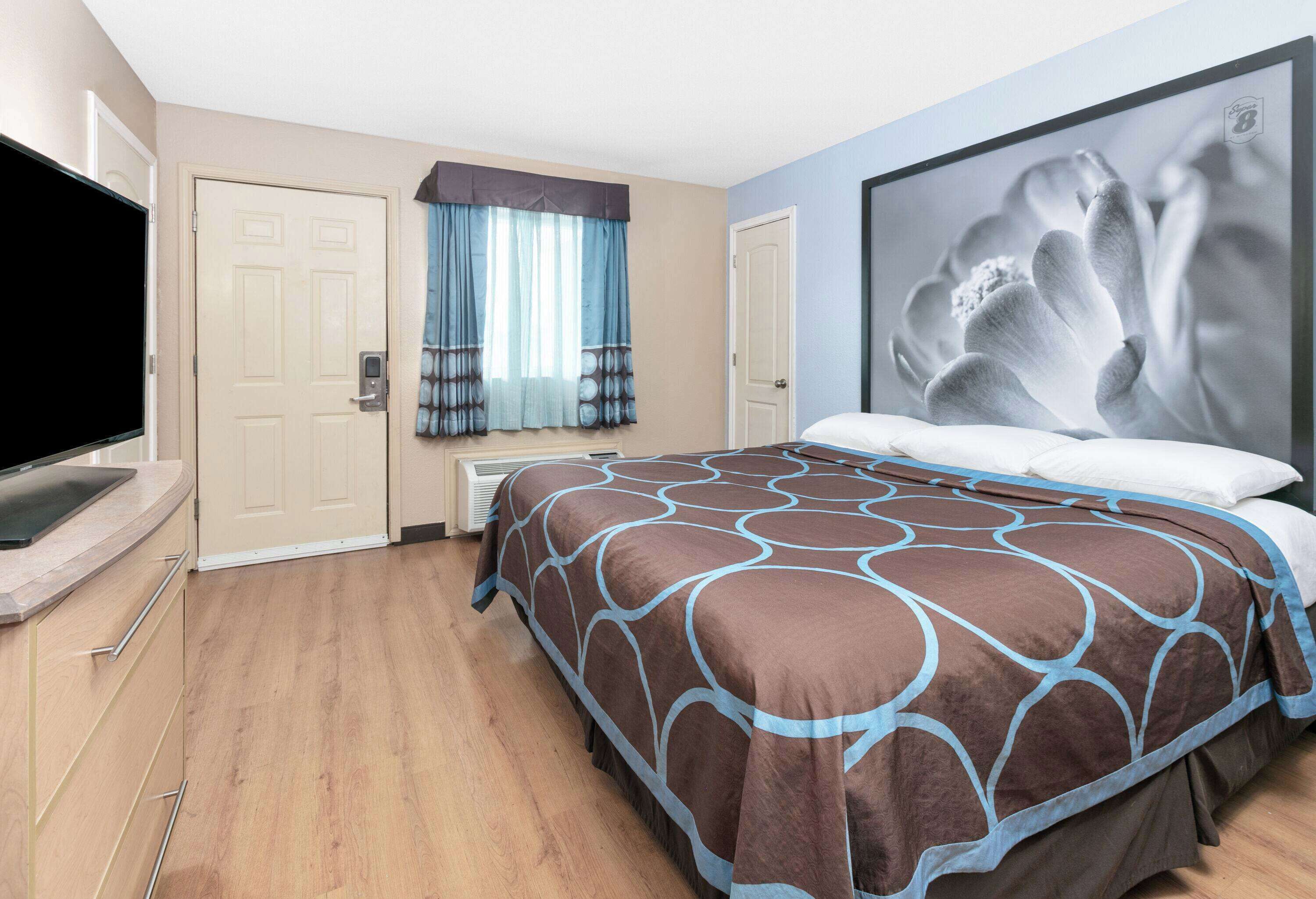 Habitación de hotel con una cama grande, un televisor de pantalla plana, una ventana con cortinas y una obra de arte colgada en la pared.