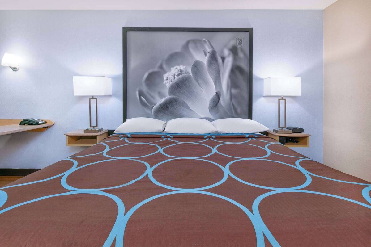 Een hotelkamer met een groot bed, twee nachtkastjes, een wandlamp en een lijst met een afbeelding boven het bed.