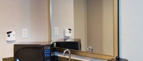 A modern bathroom with a granite countertop, a black microwave, a sink with a faucet, a mirror, and a black container labeled "E".