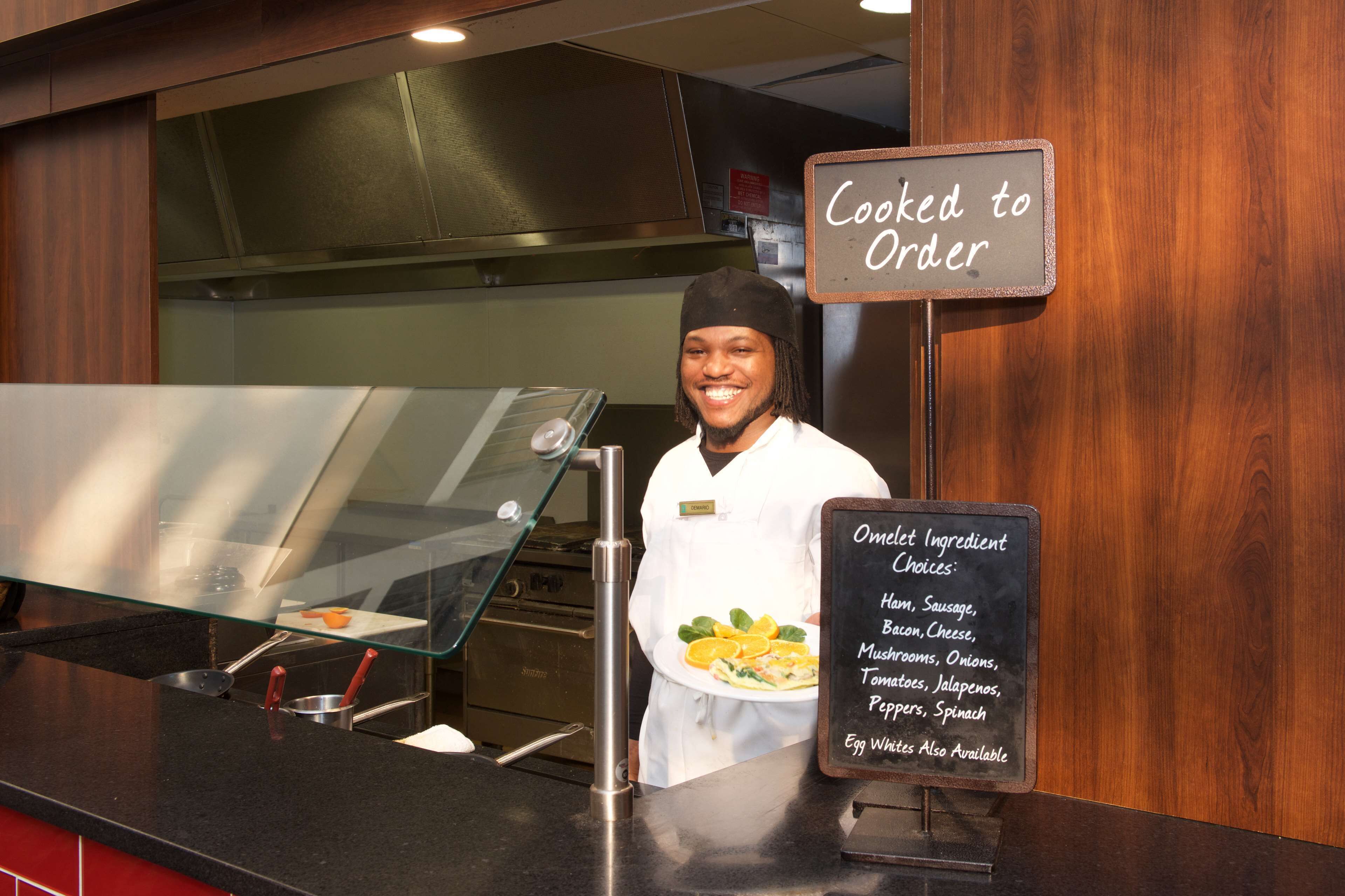 Un chef se tient derrière un comptoir, et un tableau affiche la liste des ingrédients disponibles pour les omelettes.