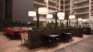 A spacious hotel lobby with multiple seating areas, including a long communal table and several smaller tables with chairs. There are large, modern pendant lights hanging from the ceiling, and the space is divided by dark wooden partitions. - Embassy Suites by Hilton Detroit Livonia Novi (Livonia)