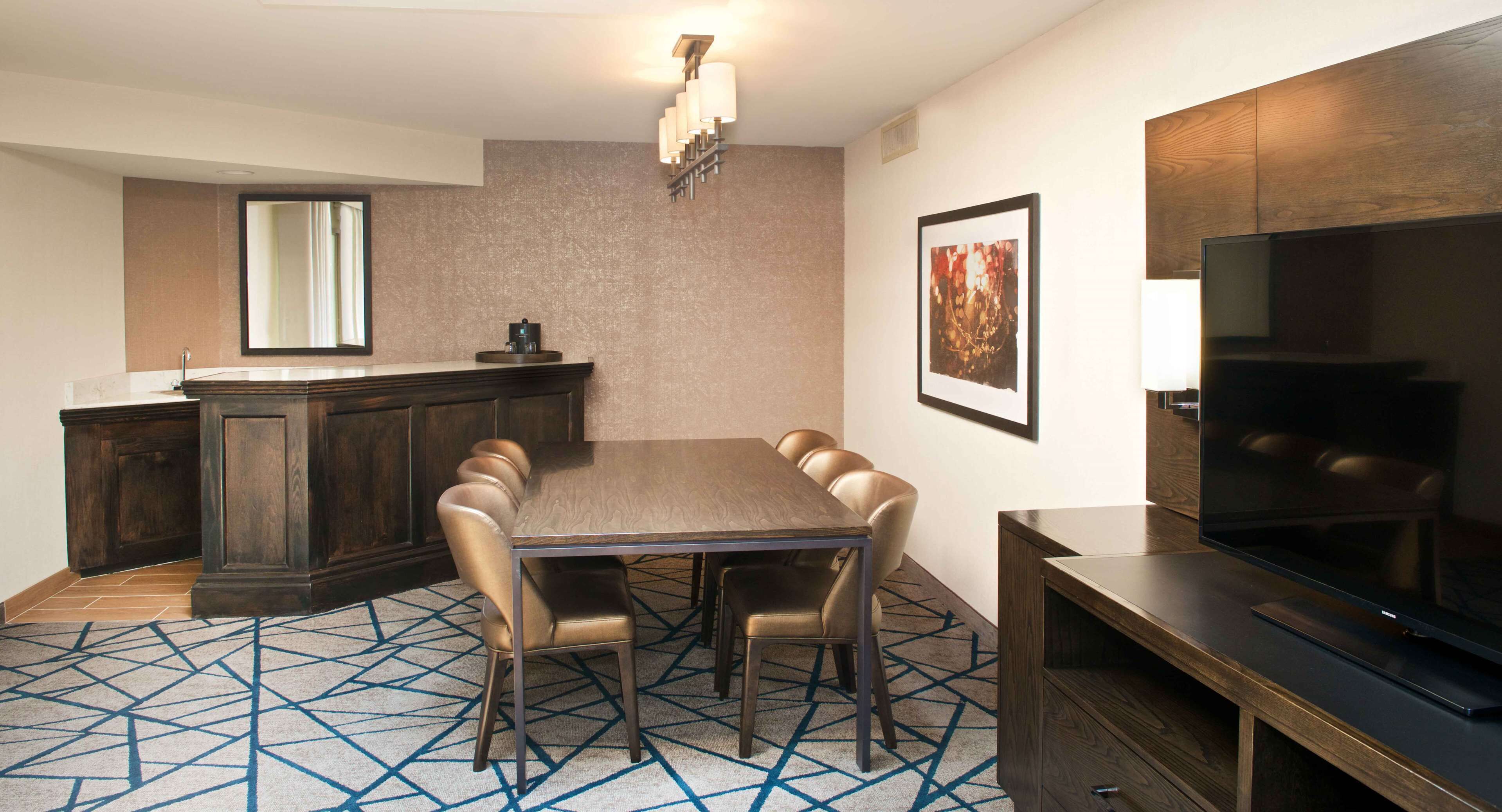 A dining area with a wooden table and chairs, a TV on a stand, a bar area with a mirror, and a patterned carpet.