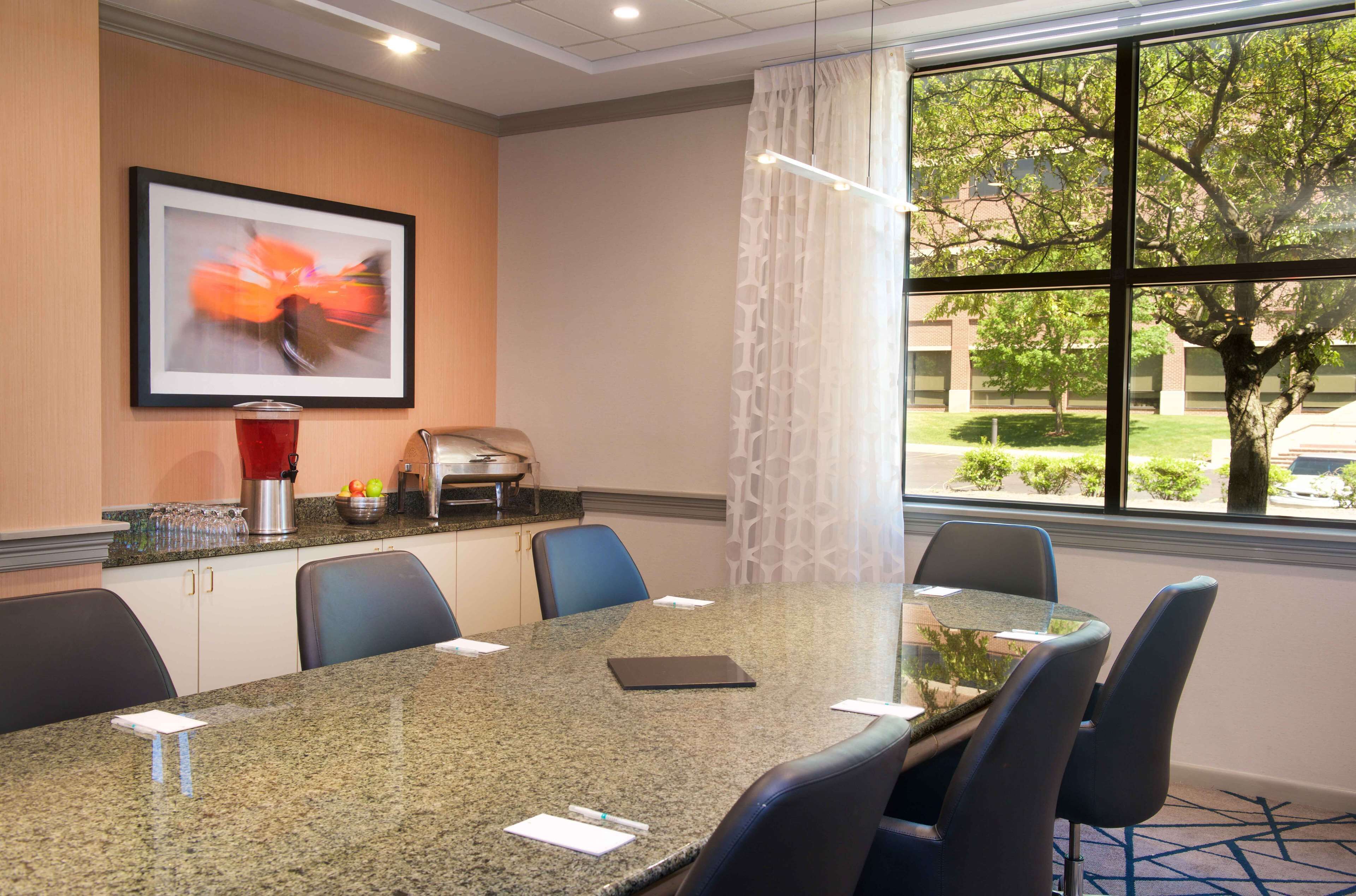 A conference room with a granite table, six chairs, a coffee station, a framed abstract painting, and a view of a tree and building through large windows.