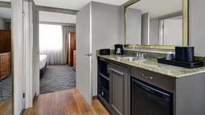 A hotel room with a kitchenette, a bed, a bathroom with a large mirror, and a view of the hallway. - Embassy Suites by Hilton Detroit Livonia Novi (Livonia)