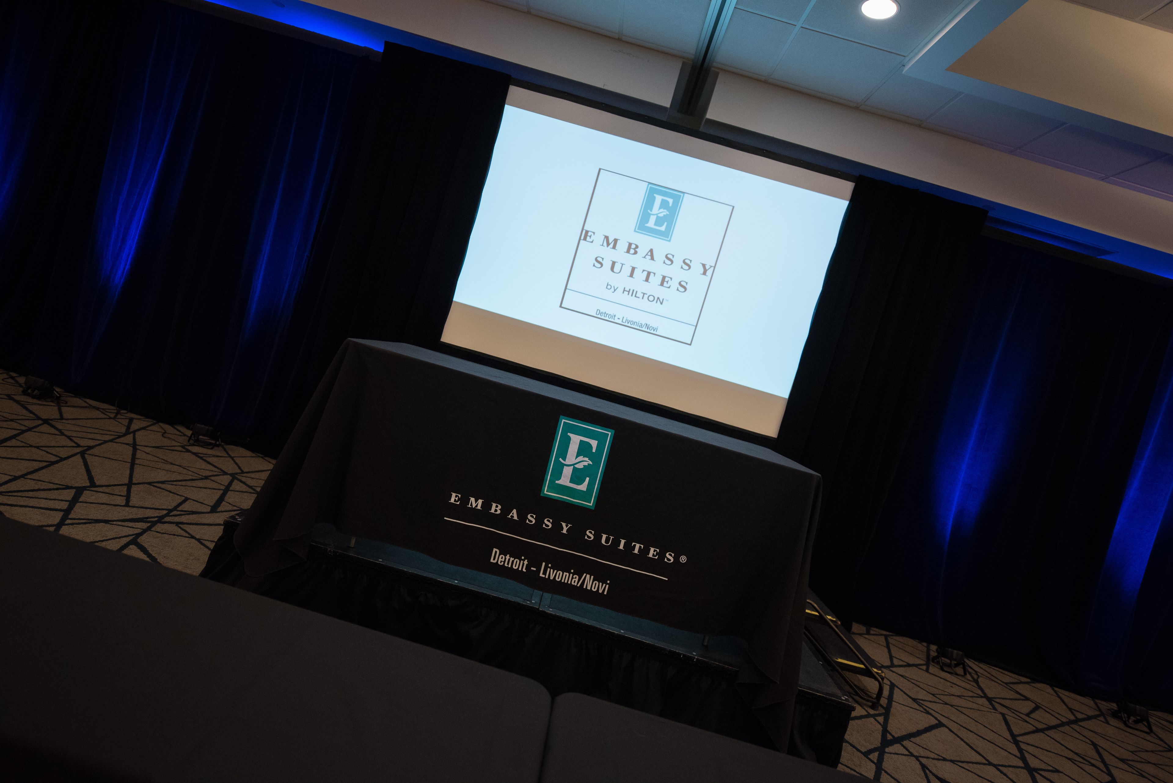 A presentation screen with the text "Embassy Suites by Hilton Detroit - Livonia/Novi" and a podium with the same logo and text.
