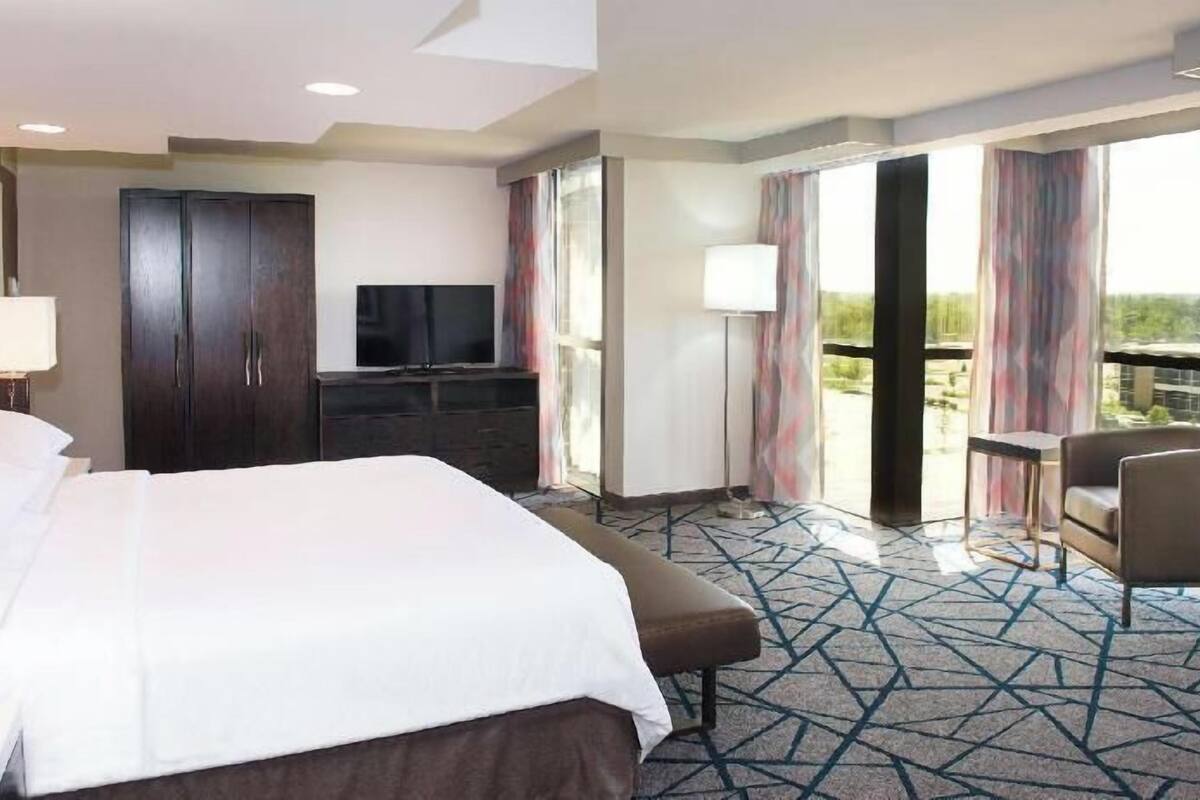Ein Hotelzimmer mit einem großen Bett, einem Fernseher, einem Schreibtisch und einem Balkon mit Vorhängen.