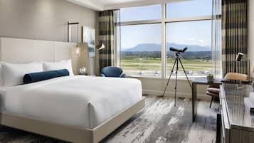 Une chambre d’hôtel avec un grand lit, un bureau avec une lampe, une chaise, un télescope et une vue sur une piste d’atterrissage et des montagnes.