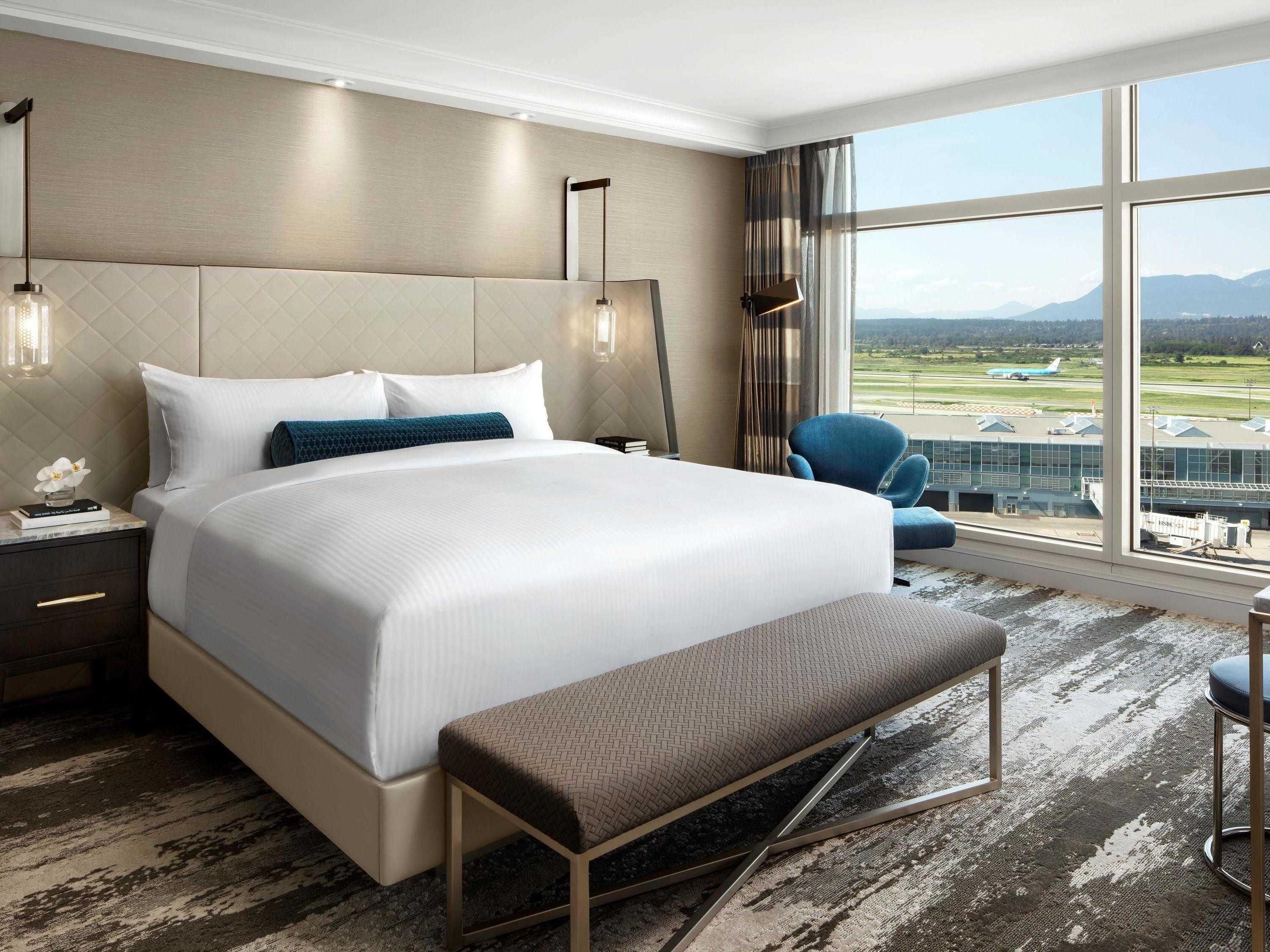 A hotel room with a large bed, a bench, a chair, a nightstand, and a view of the airport.