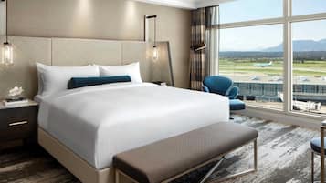 A hotel room with a large bed, a bench, a chair, a nightstand, and a view of the airport.