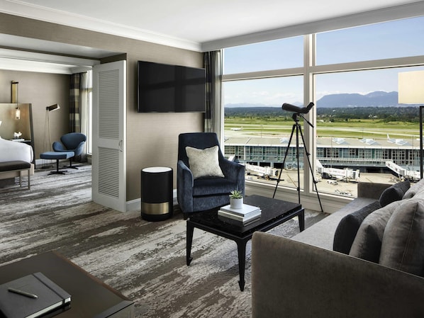 Una habitación de hotel con un ventanal grande, un televisor de pantalla plana, un escritorio con lámpara, una silla y un sofá.
