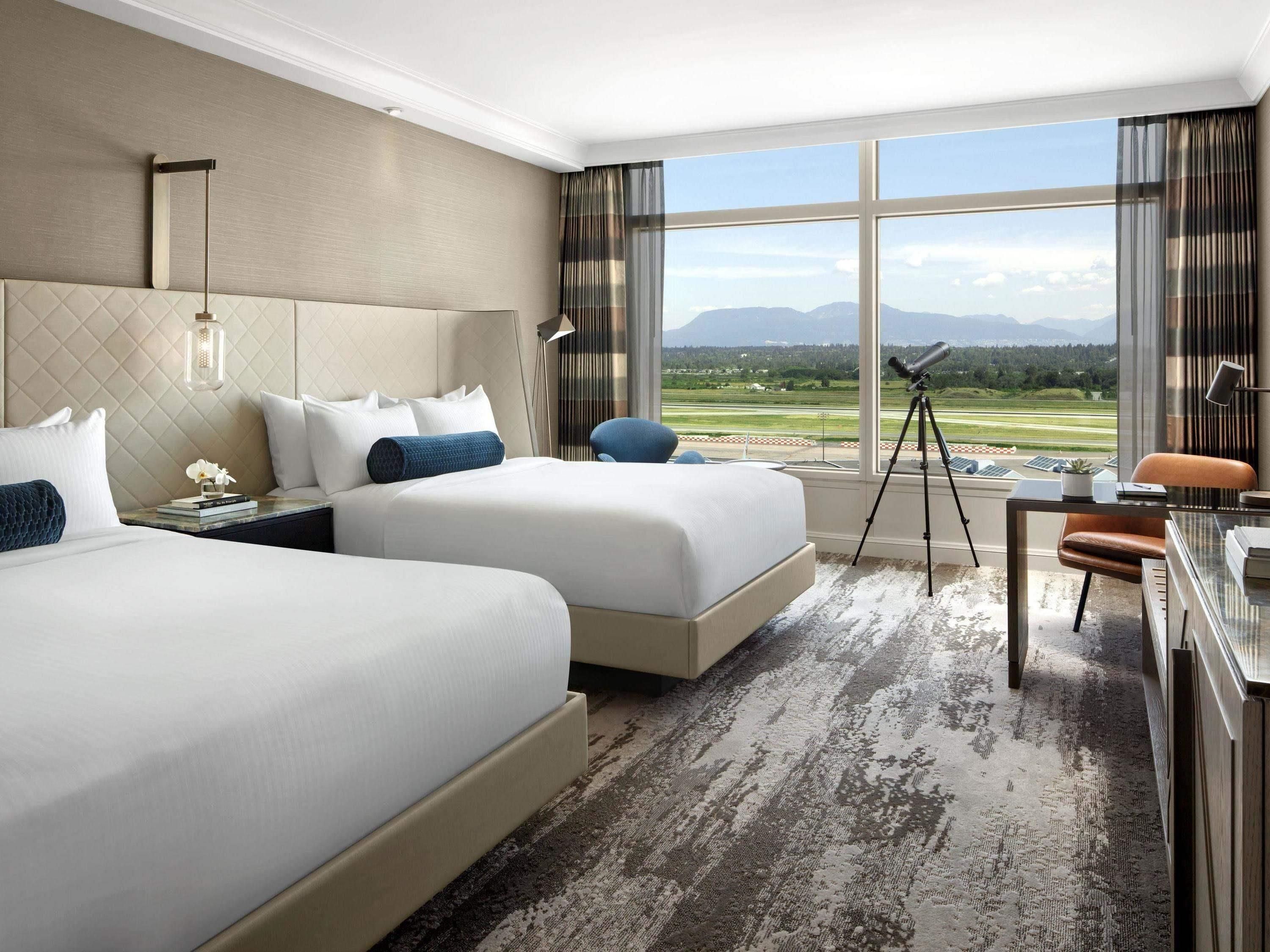 Habitación de hotel con dos camas, un escritorio, una silla, un telescopio y un ventanal con vistas a un campo de golf y montañas.