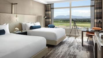Habitación de hotel con dos camas, un escritorio, una silla, un telescopio y un ventanal con vistas a un campo de golf y montañas.