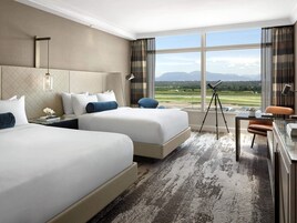 Habitación de hotel con dos camas, un escritorio, una silla, un telescopio y un ventanal con vistas a un campo de golf y montañas.