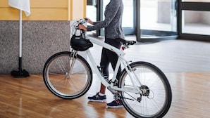 A person standing next to a white bicycle in a spacious indoor area with wooden flooring and large windows.
