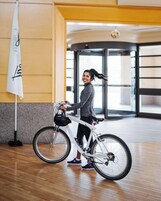 Una persona de pie junto a una bicicleta blanca en un espacio interior amplio con piso de madera y ventanas grandes.