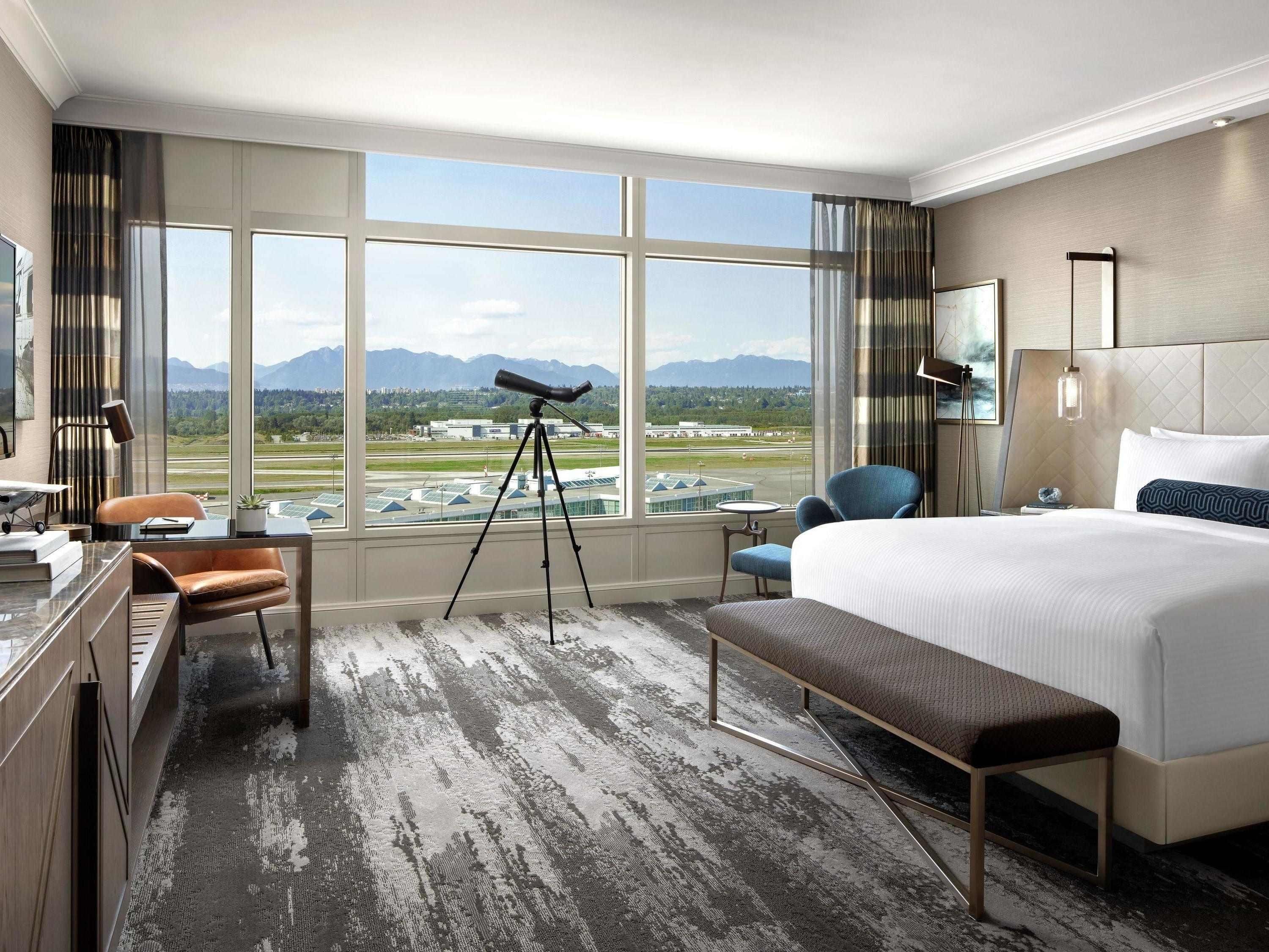 A hotel room with a large window, a telescope, a bed, a desk, and a chair.