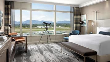 A hotel room with a large window, a telescope, a bed, a desk, and a chair.