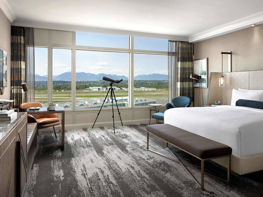 A hotel room with a large window, a telescope, a bed, a desk, and a chair.