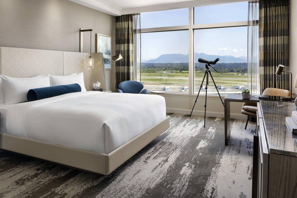 Een hotelkamer met een groot bed, een bureau met lamp, een stoel, een telescoop en uitzicht op een landingsbaan en bergen.