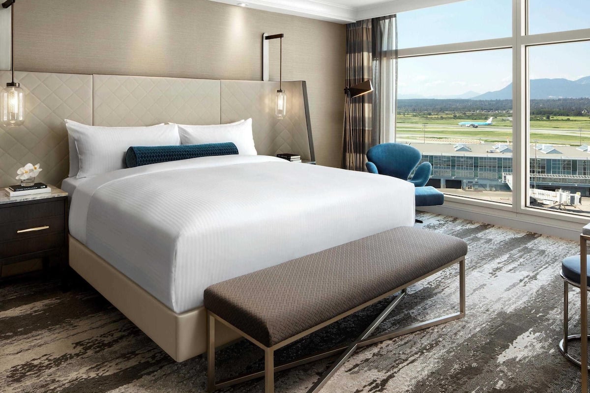 Hotelkamer met een groot bed, een bankje, een stoel, een nachtkastje en uitzicht op het vliegveld.