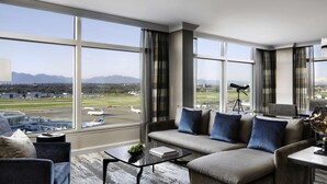 A modern hotel room with a large window offering a view of an airport and mountains.