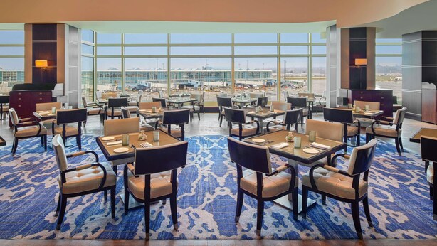 A spacious dining area with tables and chairs, large windows offering views of an airport, and a modern chandelier.