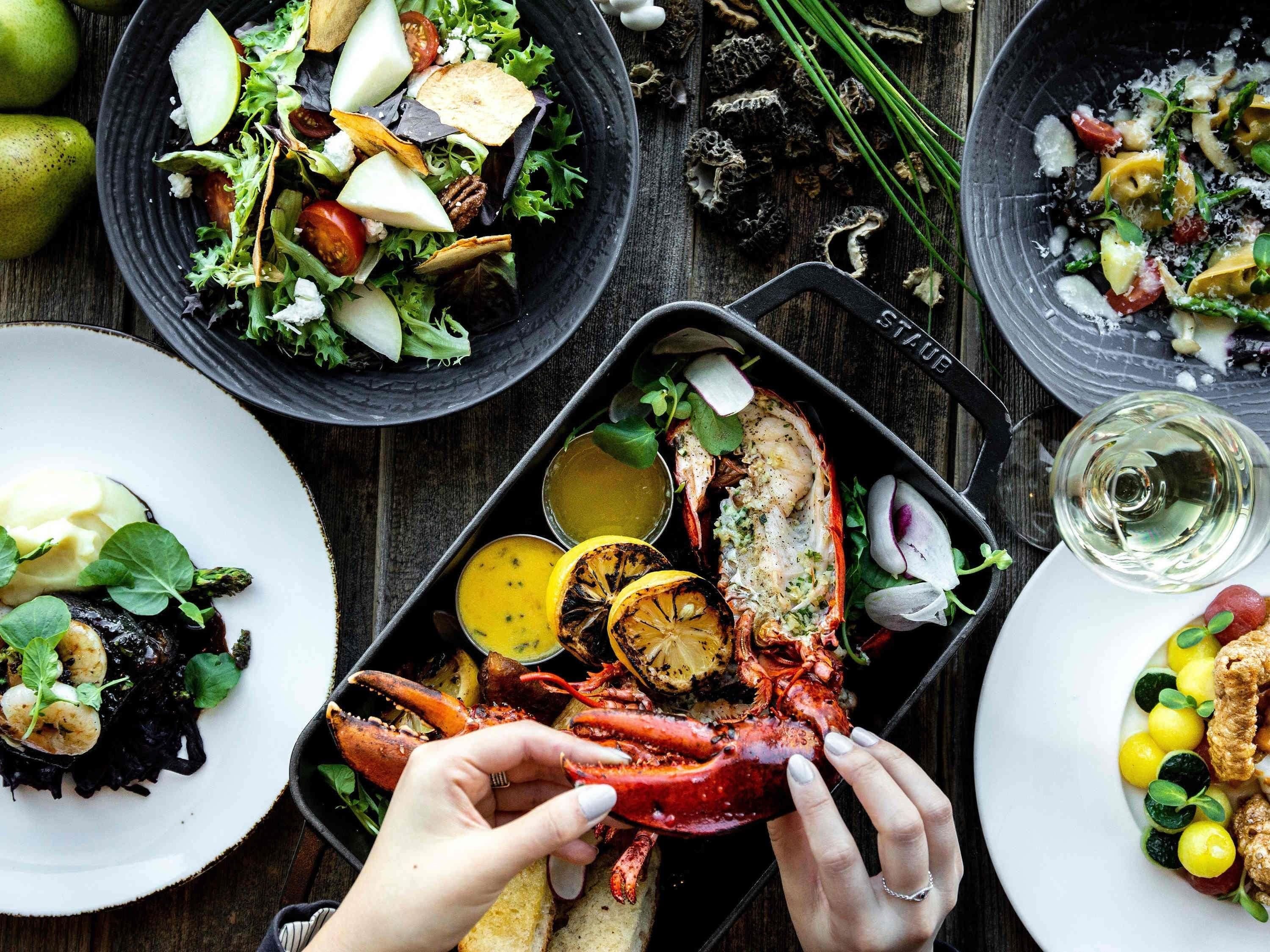 Una bandeja de mariscos con langosta, camarones y diversas verduras, acompañada de una ensalada y una copa de vino blanco.