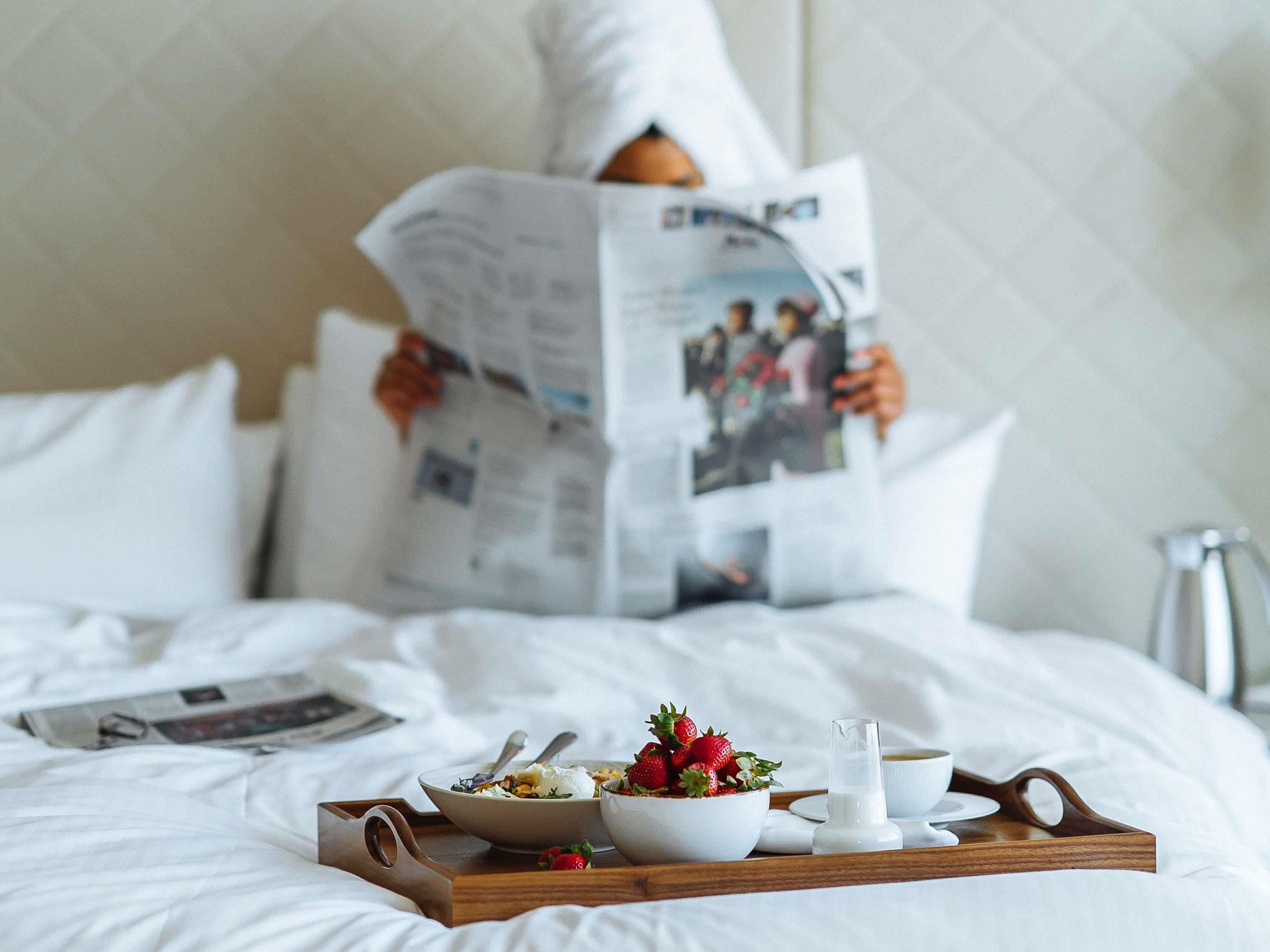 A hotel room with a bed, a tray with breakfast items, and a person reading a newspaper.