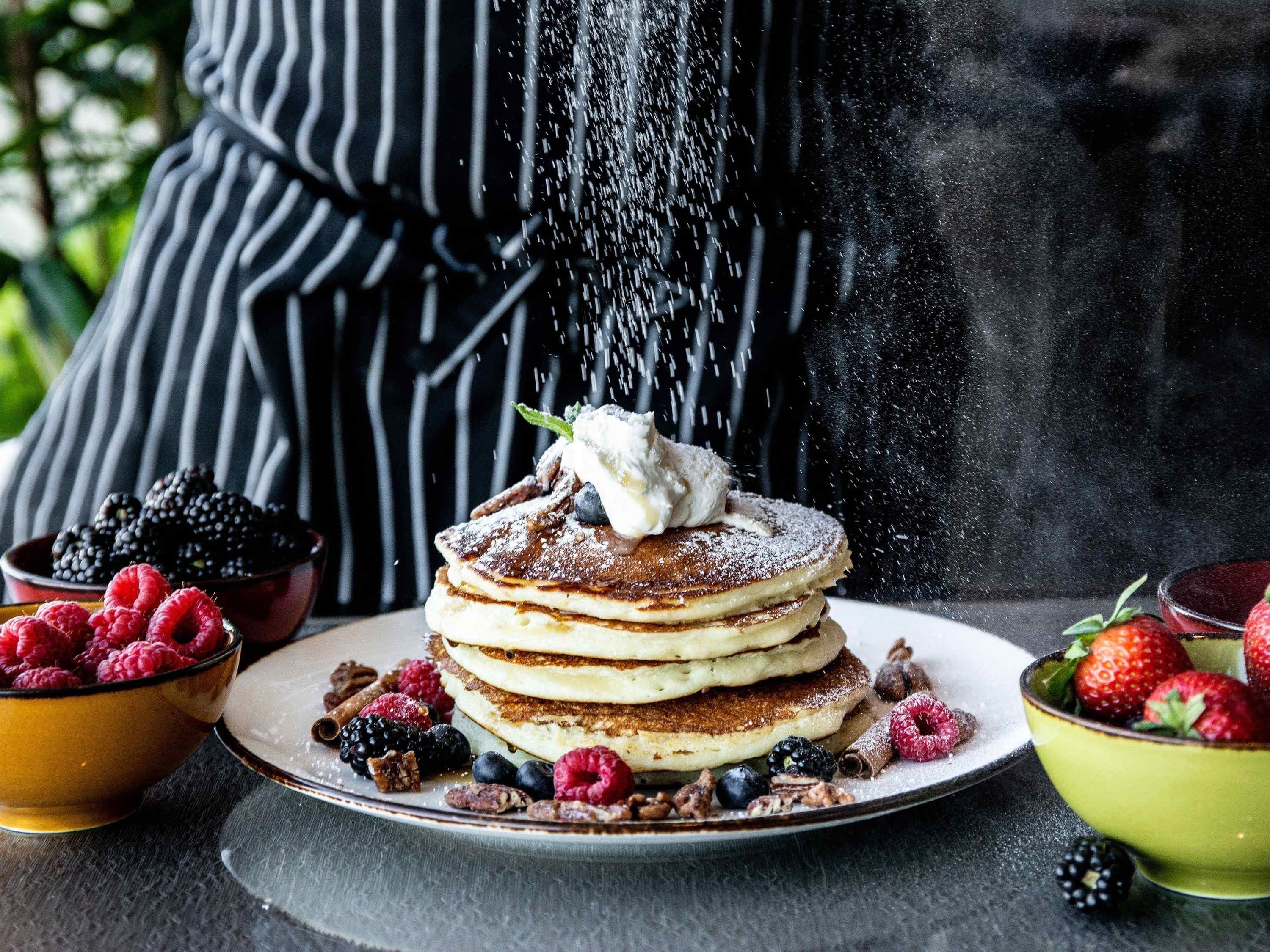 A stack of pancakes with whipped cream and berries, dusted with powdered sugar, with a person pouring sugar in the background.