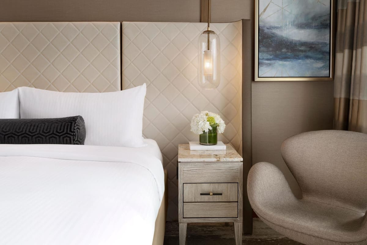 Een hotelkamer met een bed, een nachtkastje met een bloemenvaas, een stoel en een schilderij aan de muur.