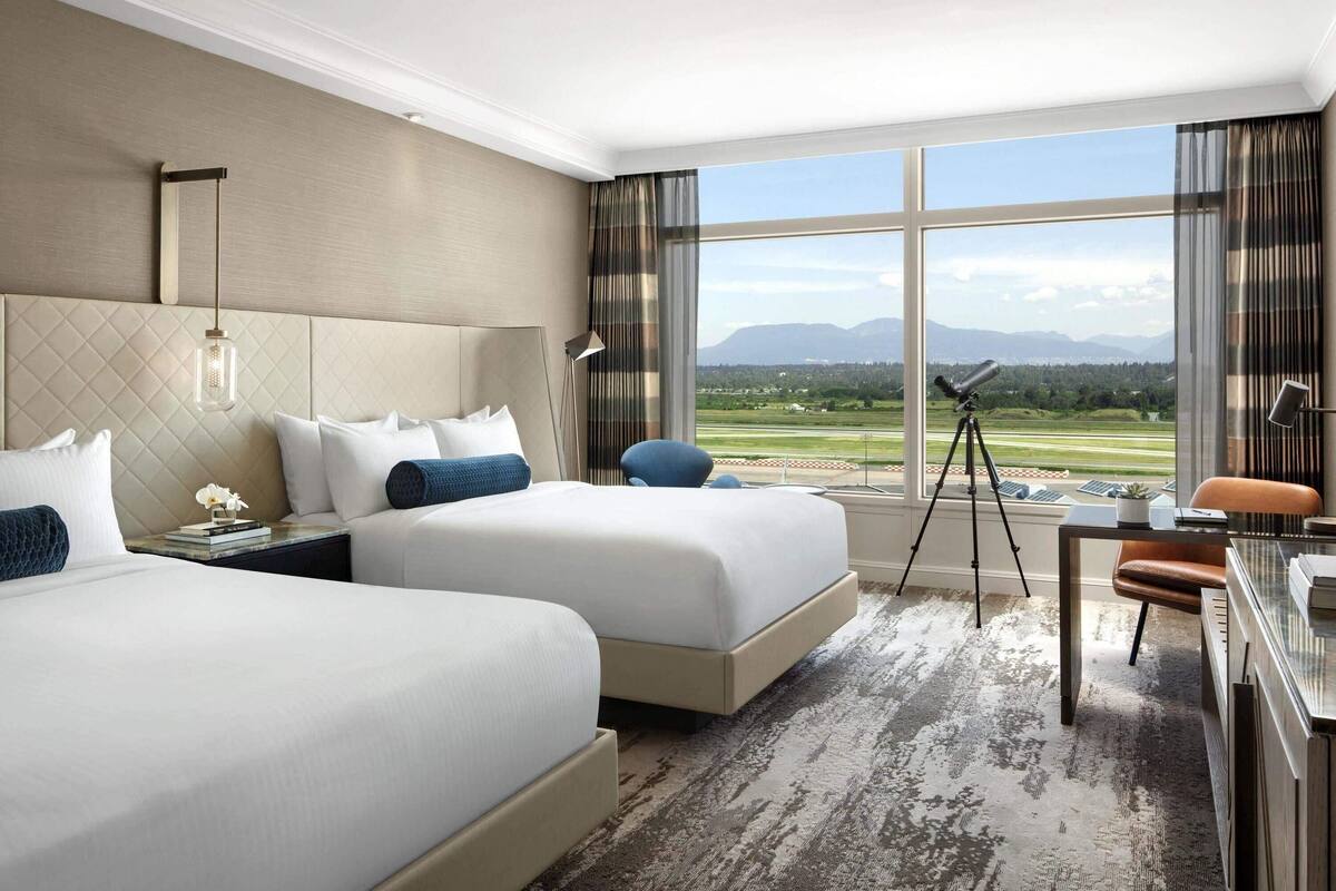 Een hotelkamer met twee bedden, een bureau, een stoel, een telescoop en een groot raam met uitzicht op een golfbaan en bergen.