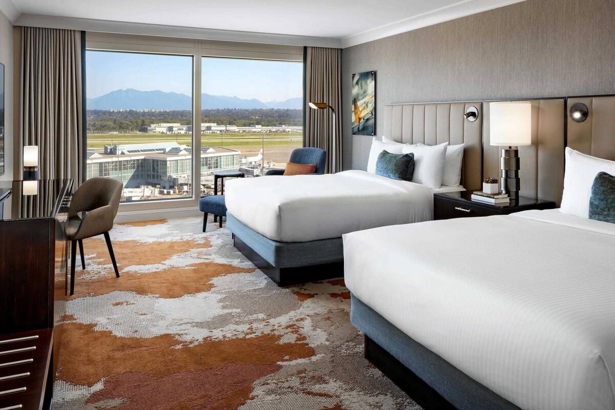 Een hotelkamer met twee bedden, een bureau en een stoel. Een groot raam biedt uitzicht op een luchthaven en bergen.
