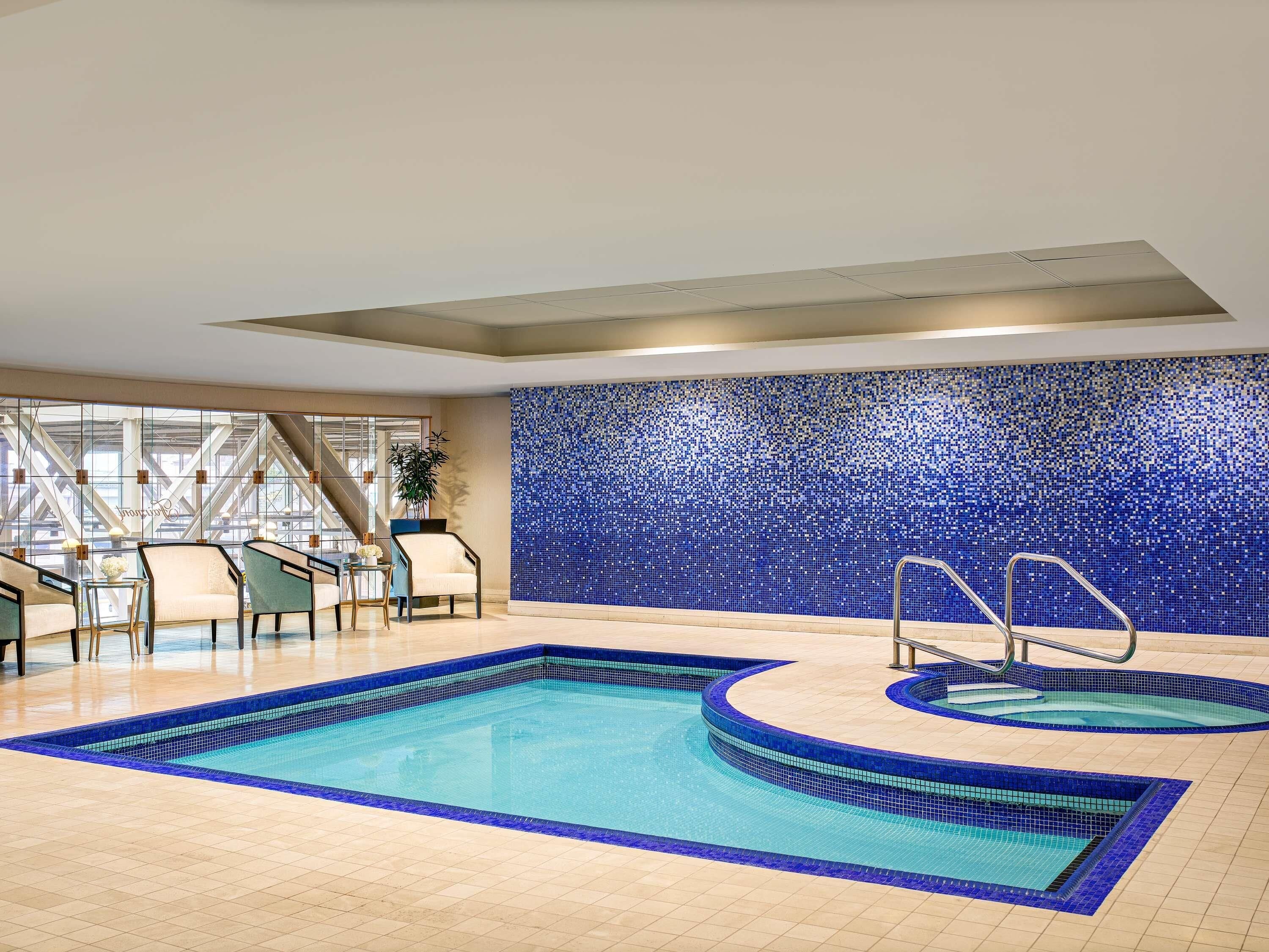 An indoor swimming pool with a blue mosaic wall, a seating area with chairs, and a staircase.