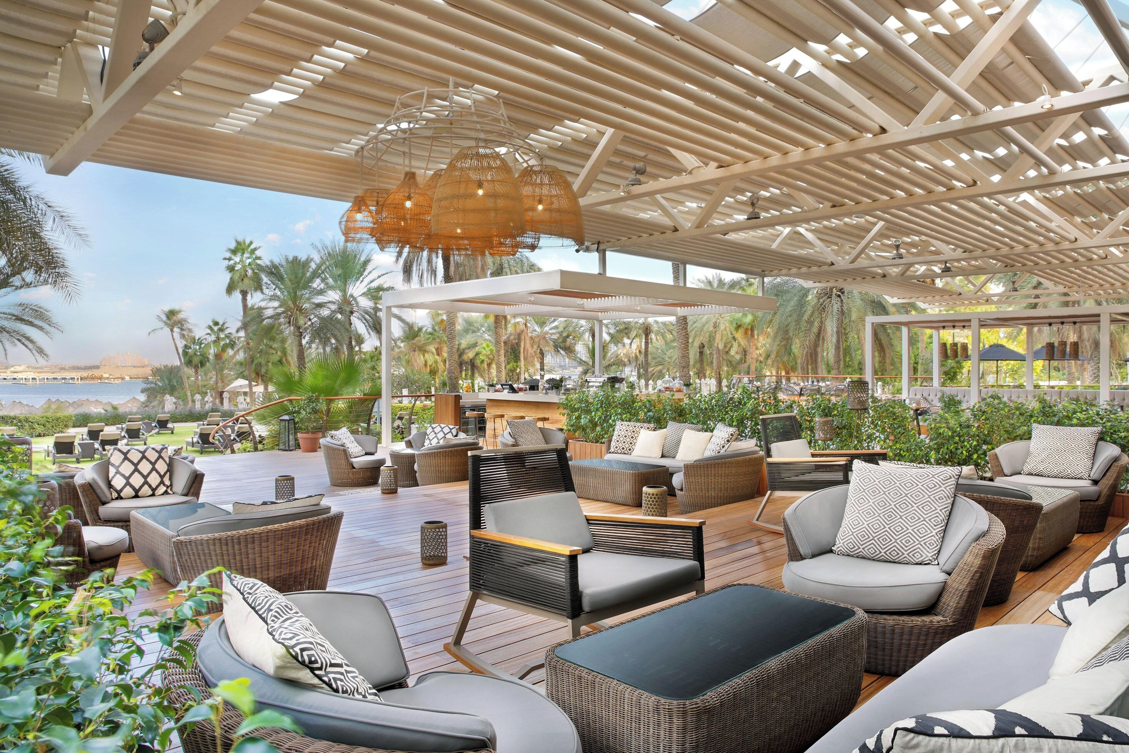 An outdoor lounge area with wicker furniture, a wooden deck, and a view of palm trees and a body of water.