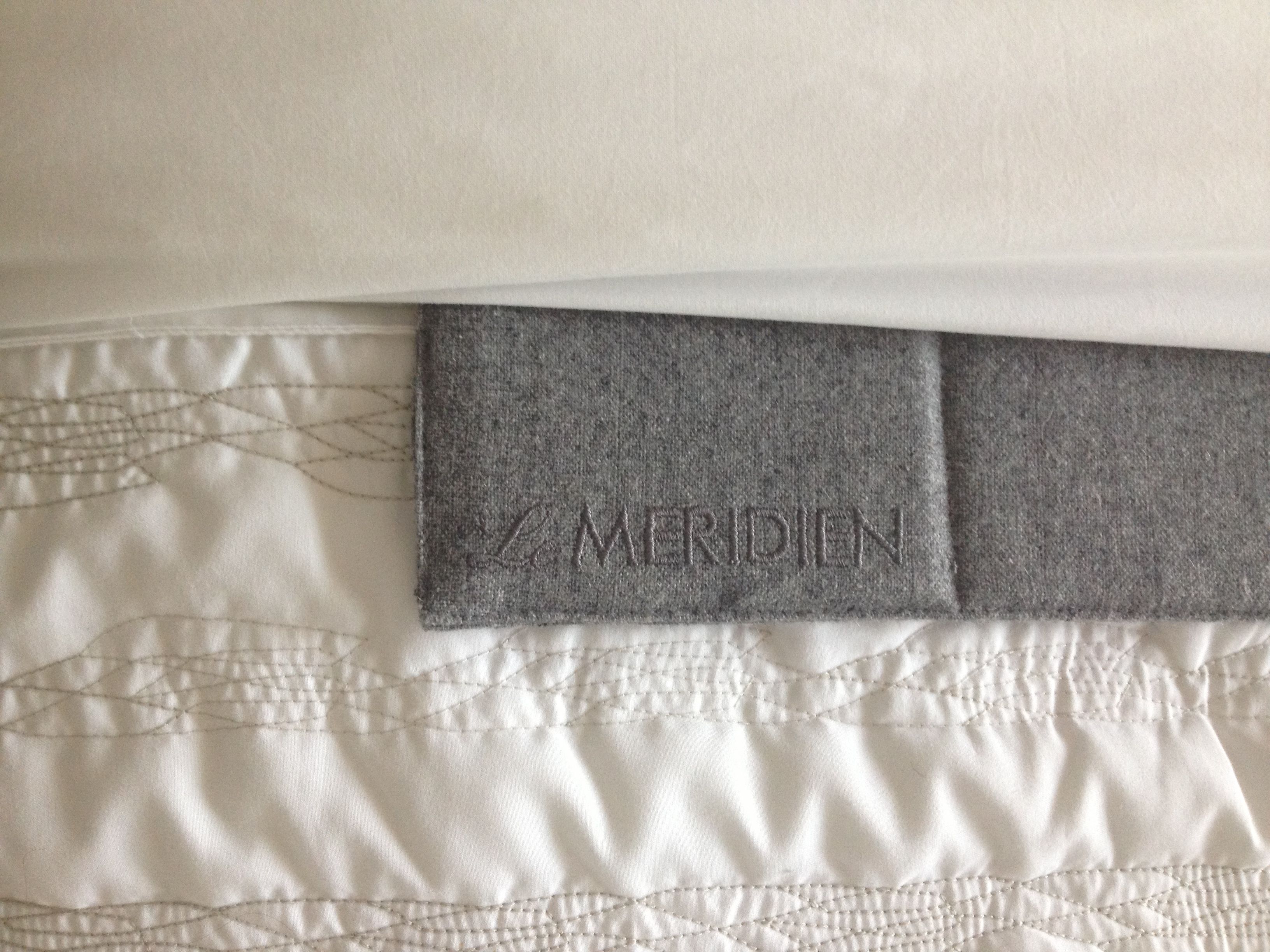 A close-up of a bed with a quilted pattern and a grey headboard with the brand name "MERIDIEN" embossed on it.