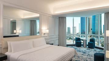 A hotel room with a large bed, a mirror, a view of tall buildings, and a balcony with chairs.