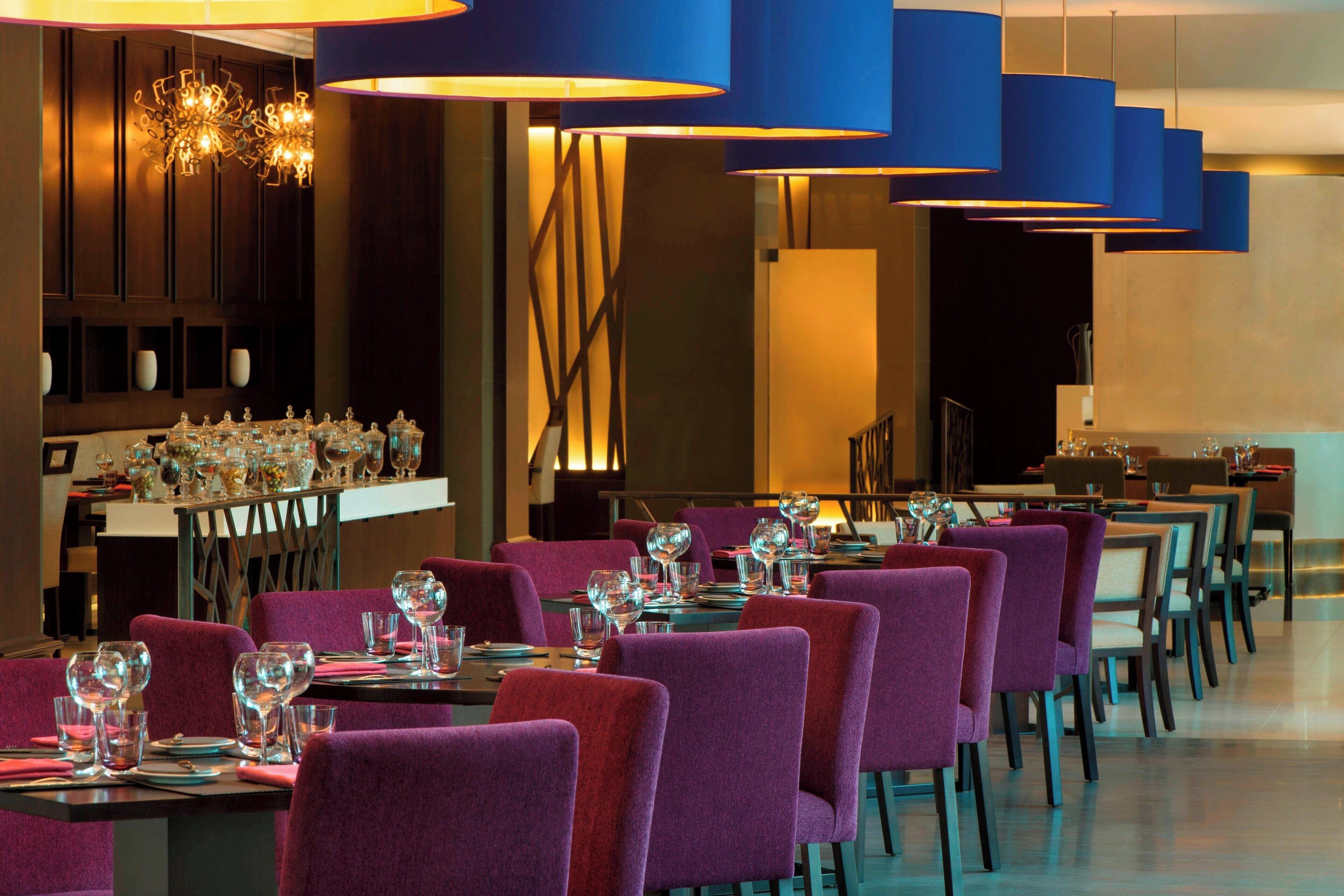 Een restaurant met paarse stoelen, gedekte tafels met wijnglazen en blauwe hanglampen.