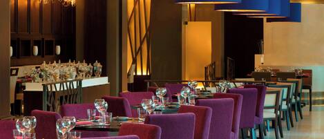 Een restaurant met paarse stoelen, gedekte tafels met wijnglazen en blauwe hanglampen.