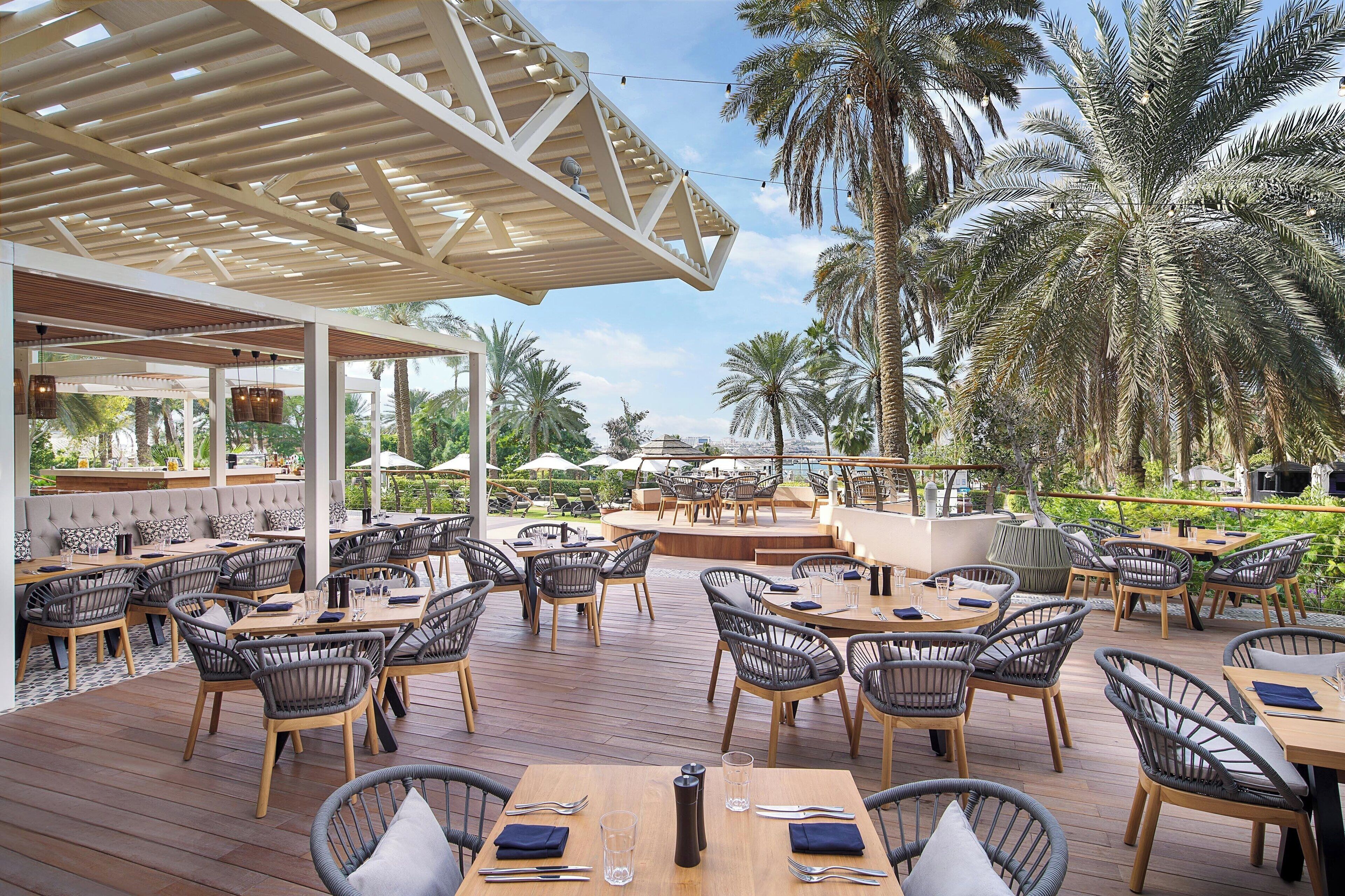 An outdoor dining area with wooden tables and wicker chairs, surrounded by palm trees and a covered seating area.