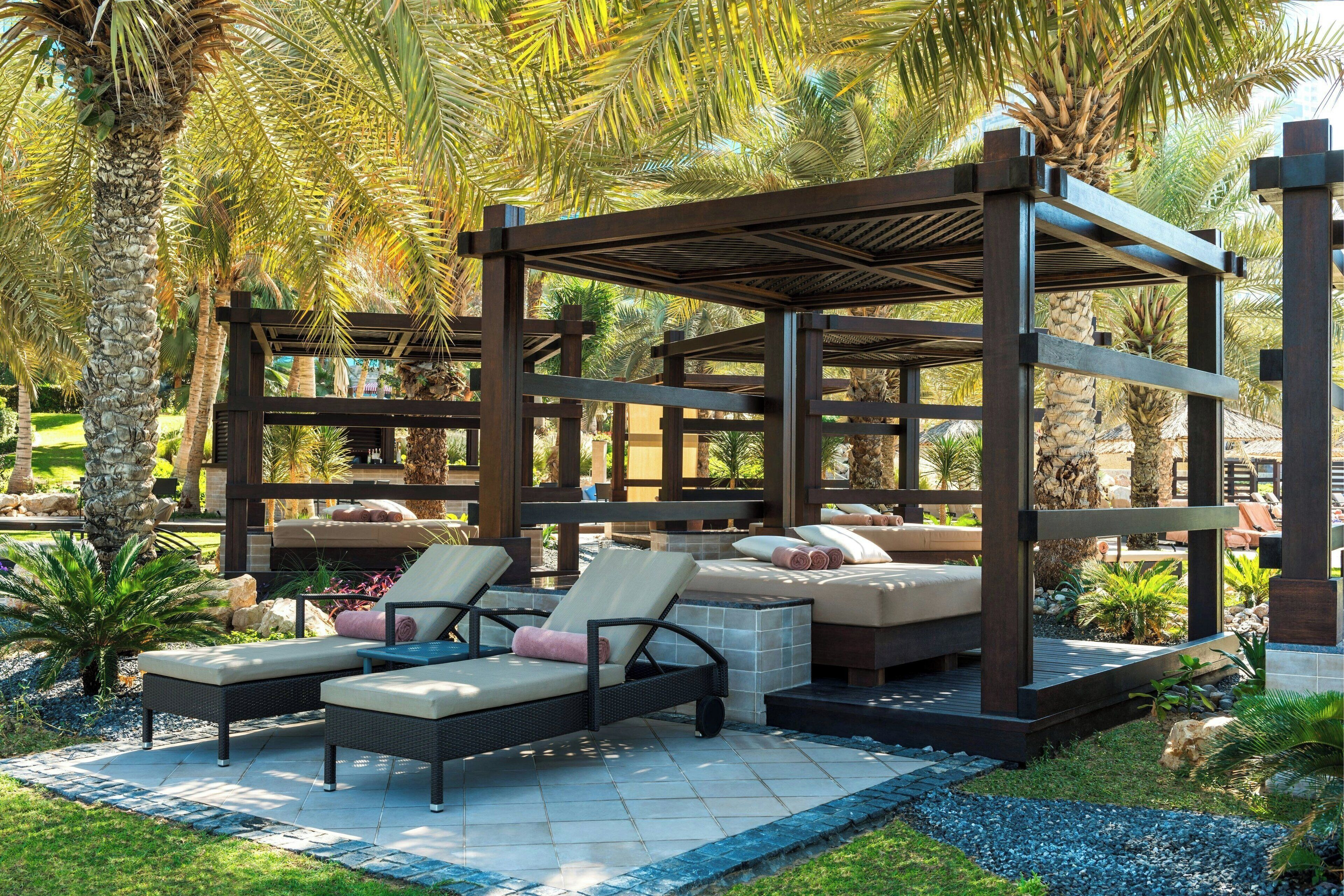 Un espace extérieur aménagé, abrité et doté de fauteuils confortables et d’un canapé intégré, entouré de palmiers et d’un jardin.