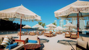 Un área de descanso junto a la playa con diversas opciones de asientos bajo grandes sombrillas tejidas, con vistas a una piscina y al horizonte urbano.