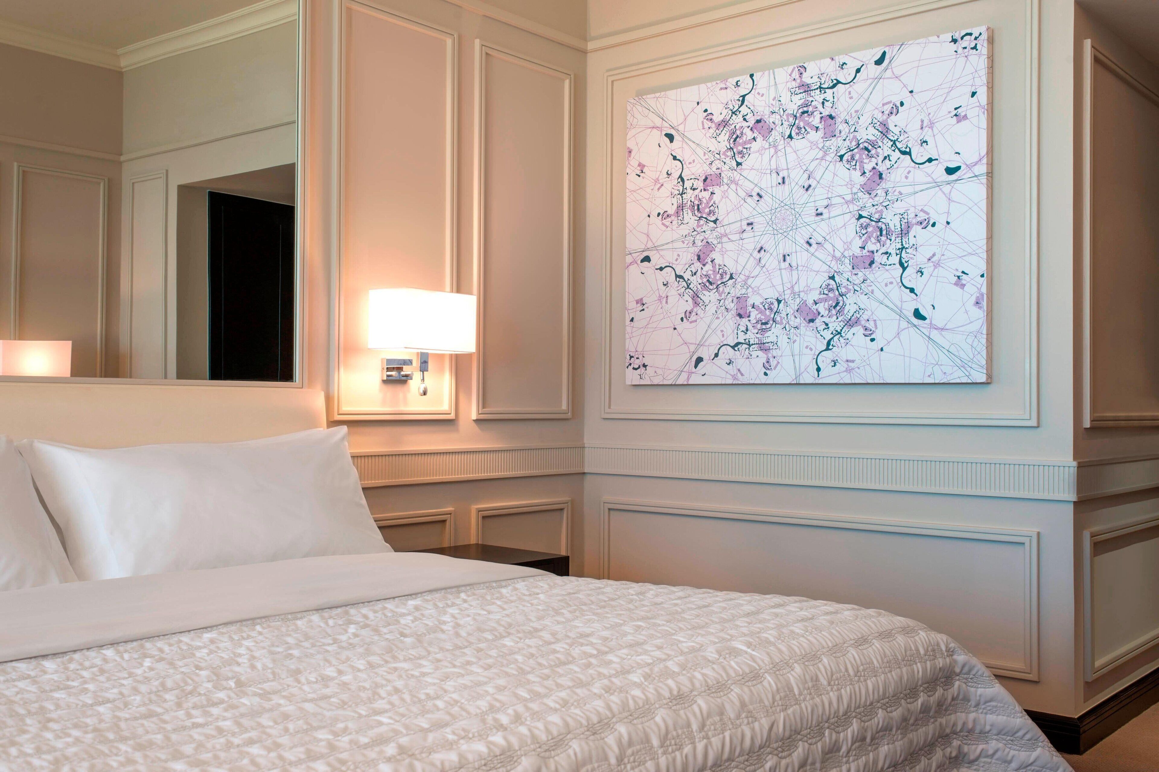 A hotel room with a large bed, a nightstand with a lamp, a wall-mounted painting, and a mirror reflecting the room.