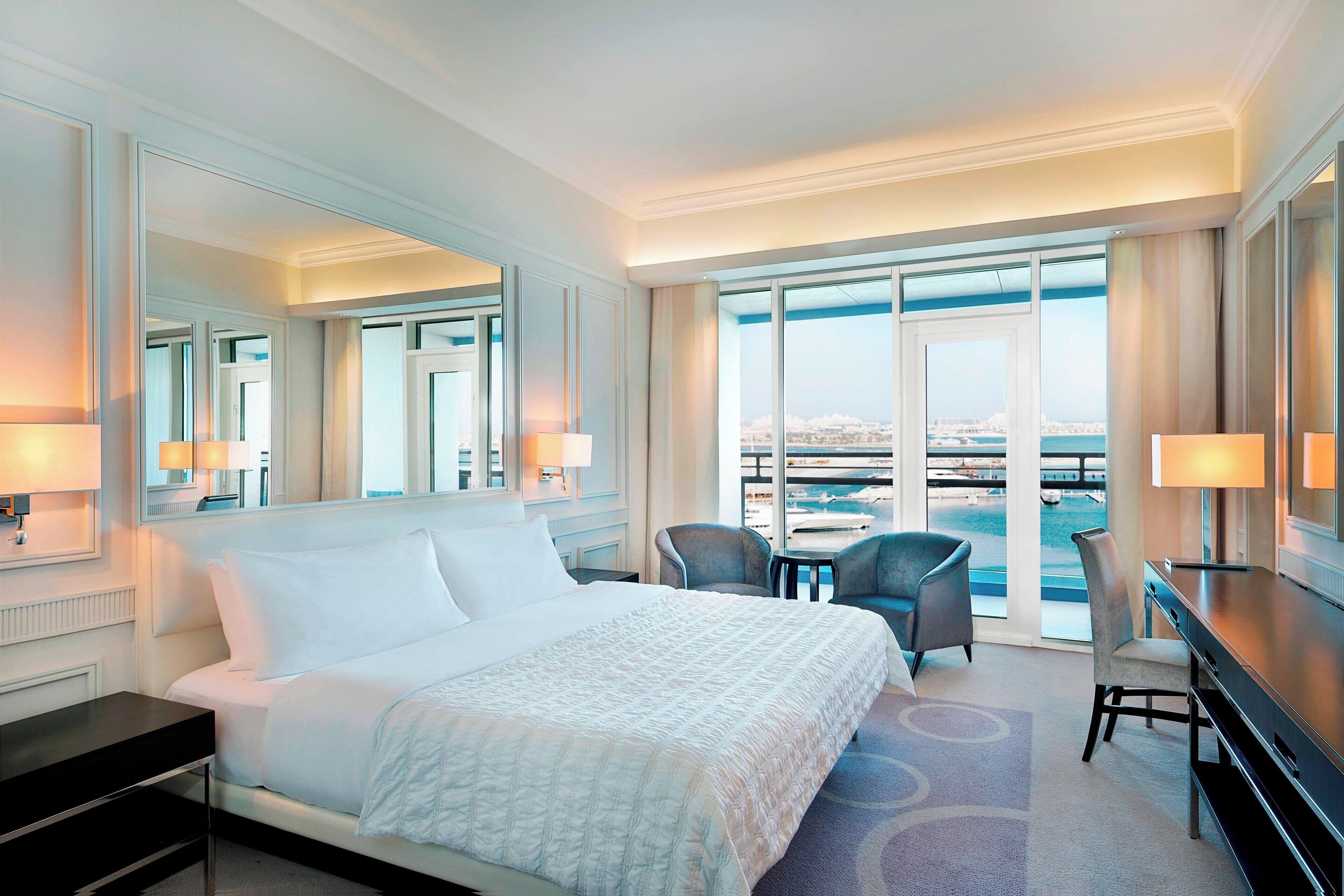 A hotel room with a large bed, a desk, two chairs, and a view of the city through large windows.