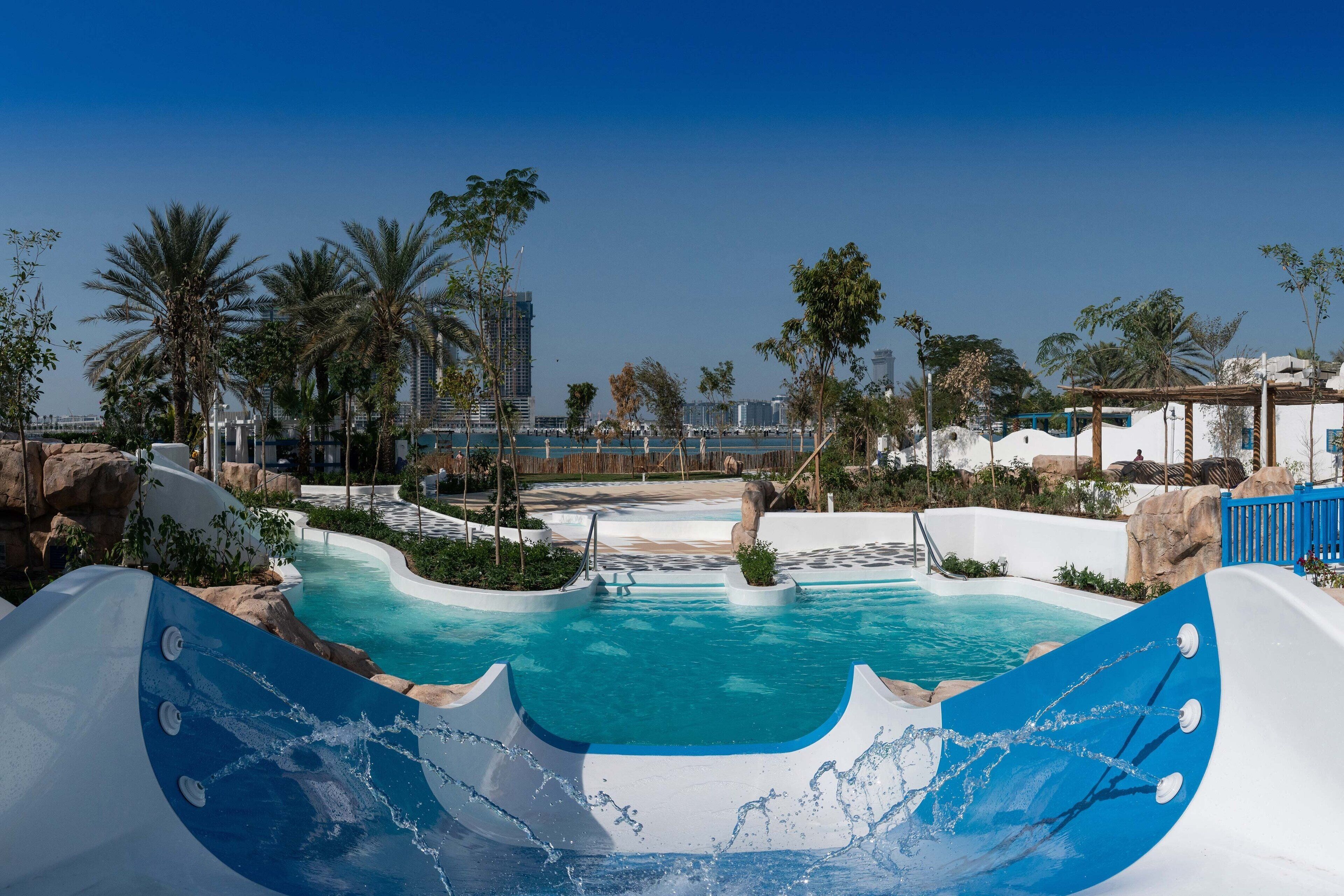 A swimming pool with a water slide, surrounded by palm trees and a view of a city skyline.